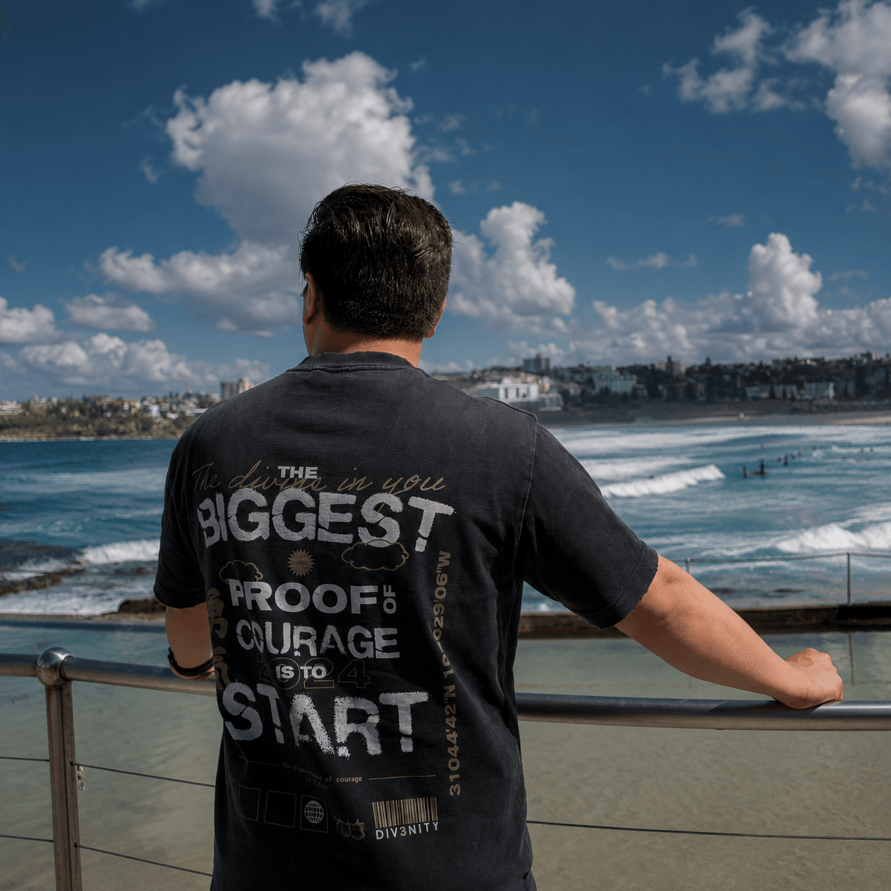 A man facing the ocean wearing the DIV3NITY BIGGEST graphic tee